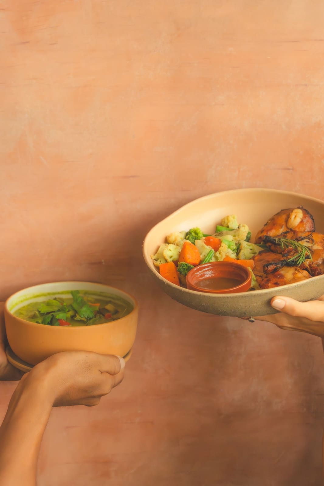 A bowl of soup and a plate of roast chicken and vegetables held against a terracotta wall.