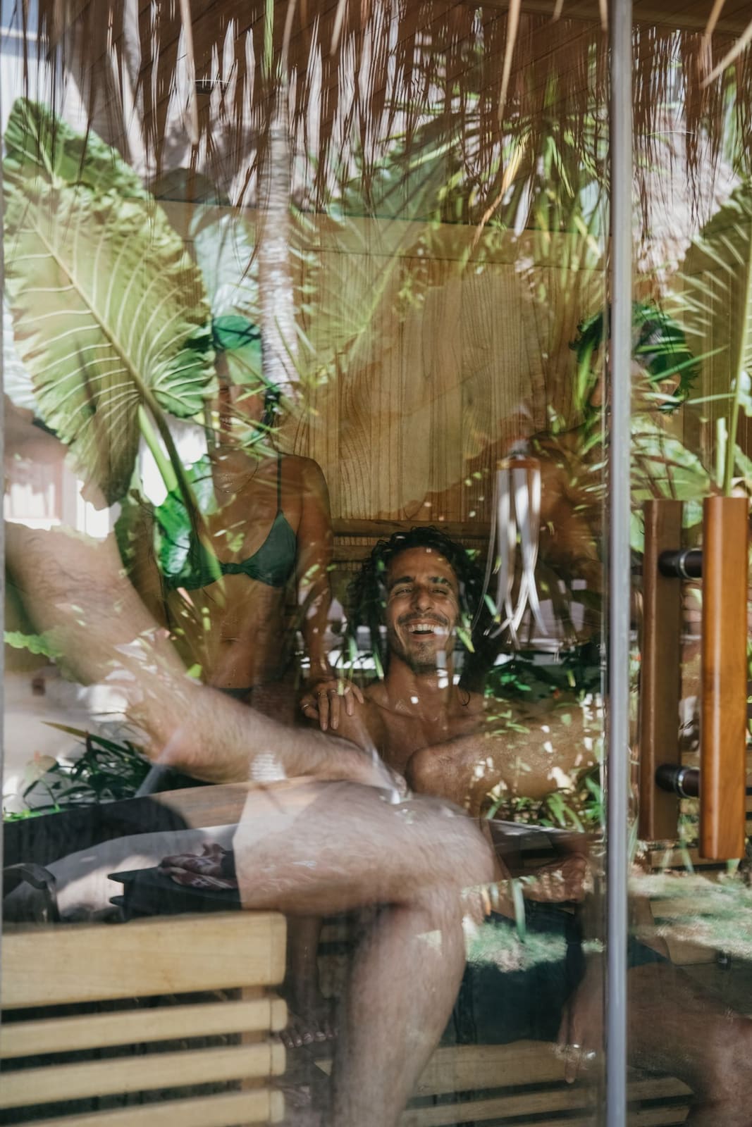Guests laughing inside the glass-fronted sauna at Lulu's, framed by palms.